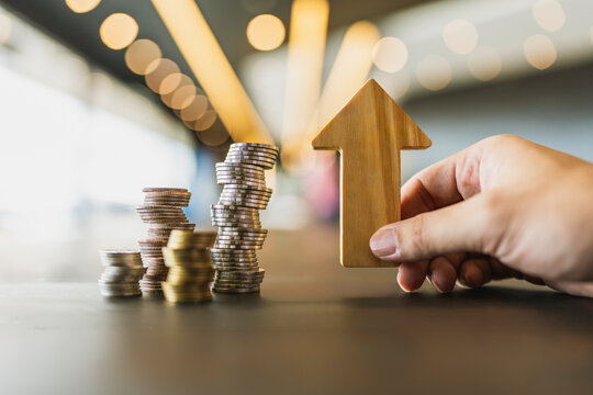 Capital appreciation and investment growth concept, hand holding wooden arrow pointing up beside ascending stacks of coins on table, representing corporate profit, revenue, and wealth rise.