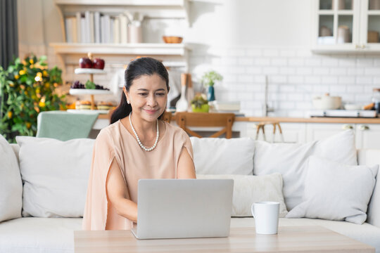 Senior digital lifestyle and remote work concept, mature Asian businesswoman with pearl necklace using laptop at home coffee table, representing active retirement, freelance, and online study.