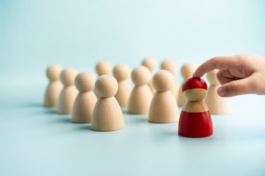 Mentorship and talent selection concept, child finger touching red wooden peg doll standing before crowd of natural wood figures on blue background, symbolizing leadership, and role model.