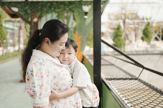 Mother and son love and family bonding concept, affectionate Asian mother in floral dress hugging young boy outdoors at park, representing maternal care, gentle parenting, and happiness.