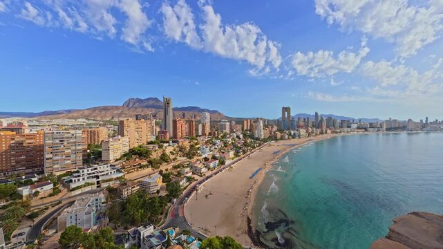 The coast of Benidorm shows buildings along the beach with mountains in the background. People walk on the sand and enjoy the clear blue water under a sunny sky.
