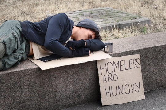 Homelessness and poverty crisis, young man sleeping on street with cardboard sign asking for help in urban environment