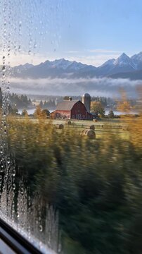 Red barn and silo slide past a wet window as hay bales, fenced fields, autumn trees, low mist, reflective water, and rugged mountains glow in soft morning light