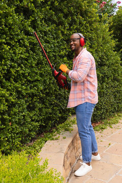 African American man trimming hedge on yard path using trimmer wearing orange gloves, copy space