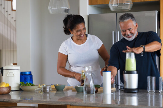 African American couple smiling while preparing green smoothie at kitchen island with blender