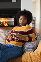 Fototapeta premium African American woman sitting at home on sofa wearing striped sweater holding glass mug near fire
