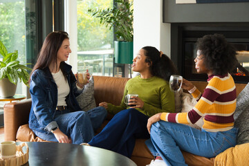 Fototapeta premium Diverse female friends sitting closely on leather sofa in modern living room holding mugs