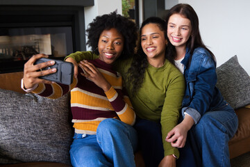 Fototapeta premium Diverse female friends sitting on sofa in living room taking selfie with smartphone case and jeans
