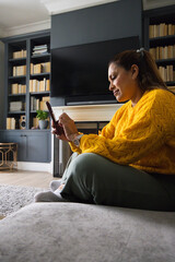 Fototapeta premium Mature woman sitting on low couch in living room wearing mustard-yellow sweater holding smartphone