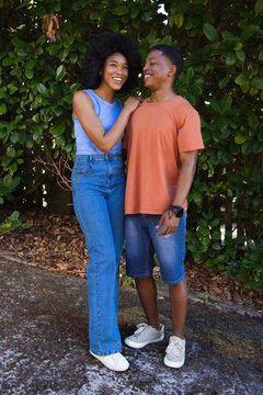 African American couple standing close, smiling and locking gazes by hedge in denim shoes, watch