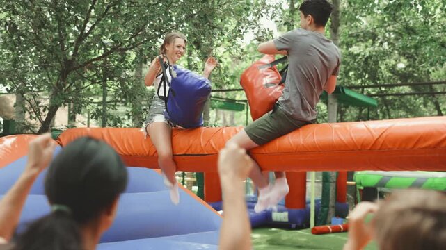 European guy and a young woman fight on bags on a log. Group of young people having fun on amusement park rides . High quality 4k footage