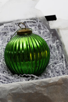 A vibrant, ribbed green glass Christmas ornament sits nestled in white crinkled paper shreds inside a gift box