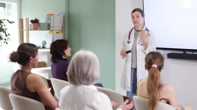 Confident adult woman doctor dressed in white medical coat with stethoscope standing and talking in auditorium where sitting medical interns different ages 