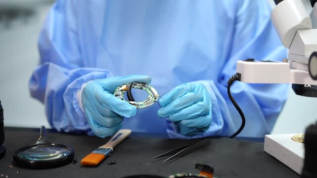 Technician in protective gown and gloves repairing a wristwatch movement at a workbench with microscope, tools and magnifying glass