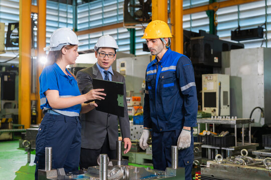 Team of industrial engineers and workers discussing project in manufacturing plant, manager and factory workers wearing safety helmets reviewing work and production quality on clipboard in workshop