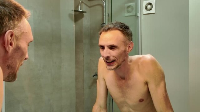 Happy athletic man washing his face and smiling at his reflection in the bathroom mirror