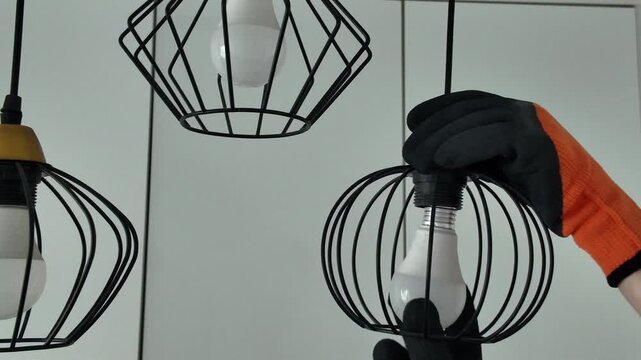 Professional electrician wearing gloves screwing a new energy-saving led light bulb into a modern black wire-frame ceiling lamp fixture