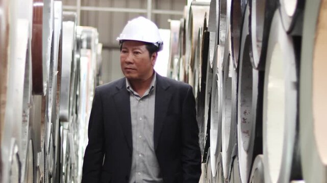 Asian senior manager in suit with helmet walking around to check large metal tube product at manufacturing plant factory. Owner businessman inspecting product and working area at industrial site.