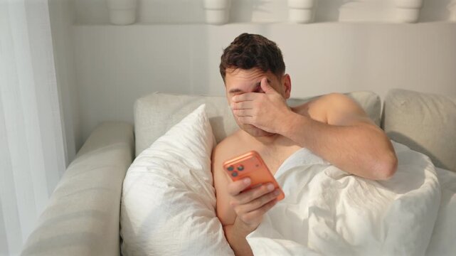 A shirtless man covers his eyes with his hand while reading an orange phone, then leans back on the pillow with closed eyes and sighs. Sofa bed, white duvet, potted plants on a shelf, morning sun.