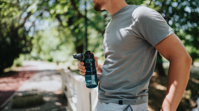 Young man holding a water bottle stands by a park path in casual workout clothes. He pauses to rest against a fence on a sunny, tree-lined trail.
