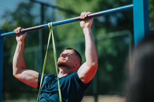 A muscular man performs a pull up using a resistance band on an outdoor bar. The athlete trains outdoors with focused effort and proper form.