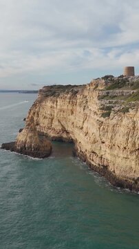 Vertical aerial footage flying along the golden coastline of Ferragudo, Portugal, passing Torre da Lapa and revealing the dramatic cliffs of the Algarve.