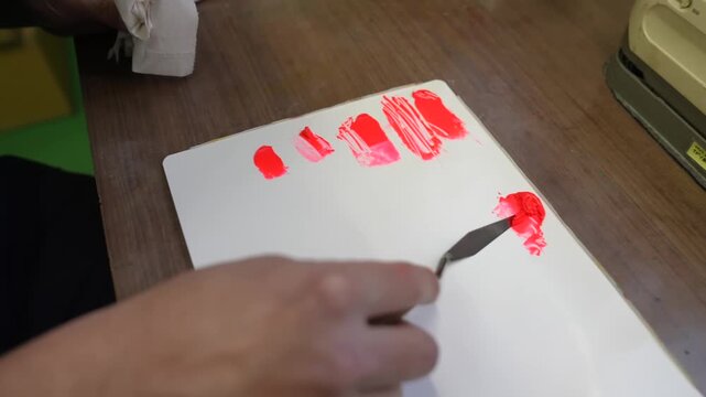 Rows of red paint swatches drawn on a white test card as a technician adds another stroke with a palette knife