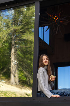 Young woman relaxing with a coffee cup by a large cabin window, peaceful cozy moment surrounded by green forest scenery and natural daylight.