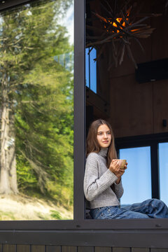 Young woman sitting by a large cabin window holding a warm coffee cup, peaceful leisure moment with green forest scenery and cozy interior.
