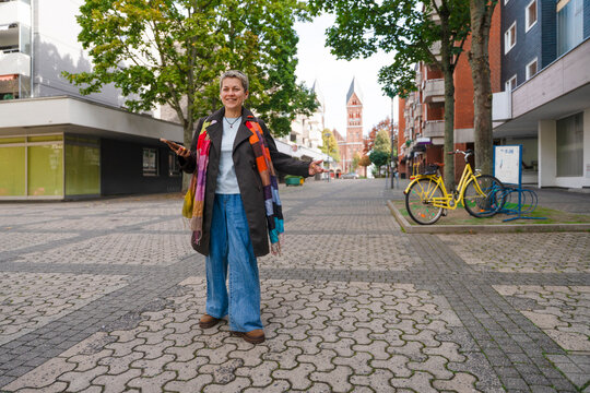 Woman stands in a public square with trees and buildings, smiling