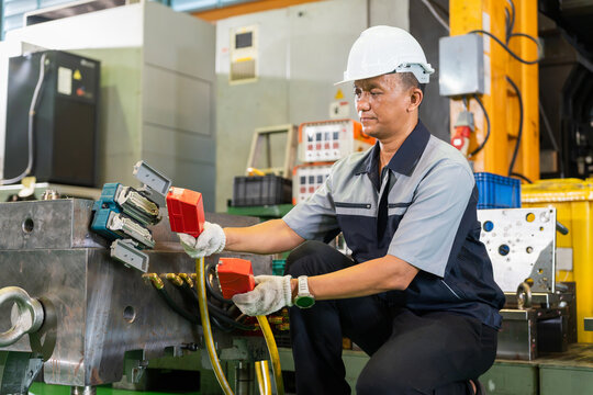 Asian factory worker wearing safety helmet connecting cable to machinery in plant, industrial technician maintaining equipment and wiring in workshop, skilled operator working on industrial machinery