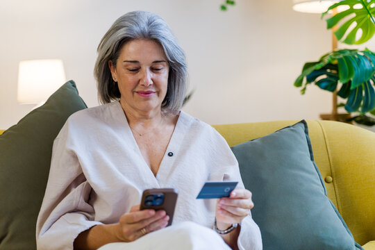 White woman using smartphone and credit card on sofa in living room