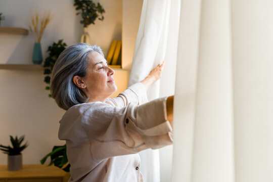 White woman opening curtains to let natural light into cozy home