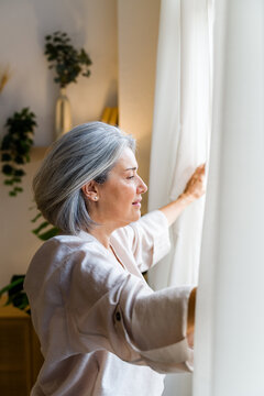 White woman opening curtain in cozy living room interior
