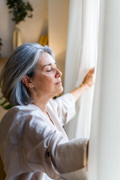Senior white woman opening curtain to natural light indoors at home