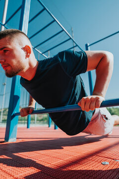 A young man performs push-ups using parallel bars during an outdoor calisthenics workout. The athlete trains upper-body strength on a sunny fitness park setup.