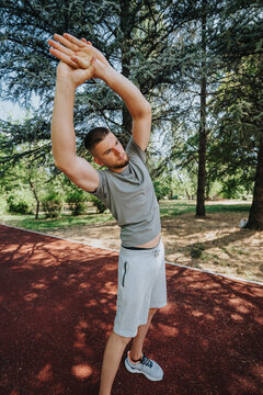 Young man stretching his arms outdoors before a run. He performs a side stretch on a red running track in a leafy park.