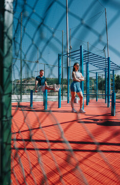 A young man and woman working out at an outdoor calisthenics park. The fitness couple train on parallel bars and ladder-style rigs in a fenced sports area.