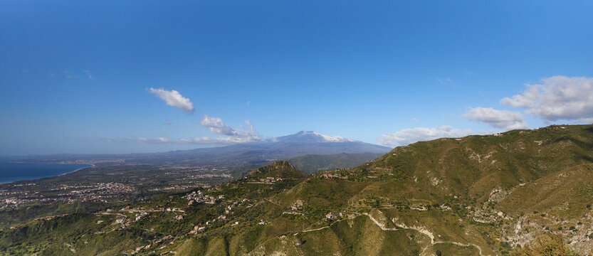 Panoramic view of  Etna volcano in Catania with a sprawling landscape under a clear blue sky, dotted with scattered clouds and green hills.