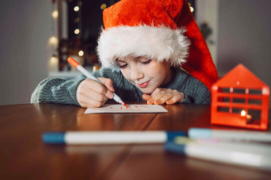 A boy in a Santa hat writes a Christmas letter indoors