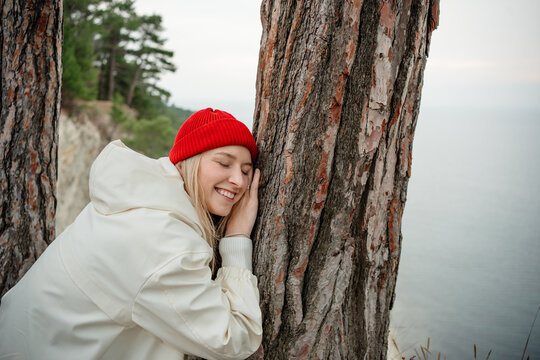 Smiling woman in a red hat hugs a pine tree on the coast