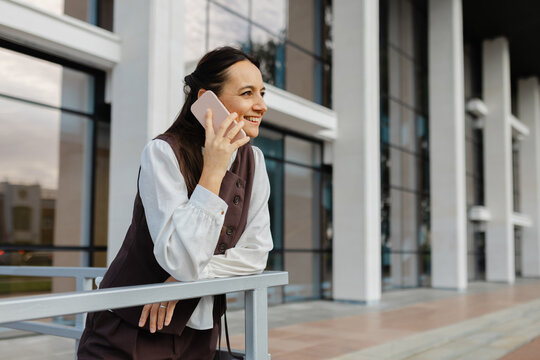 Smiling businesswoman talks on smartphone outside modern office