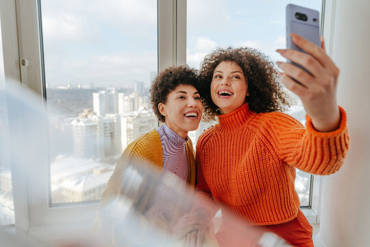 Friends with curly hair taking a cheerful selfie by a windowsill indoors