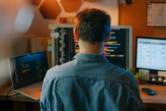 A male programmer writes code on multiple computer screens indoors