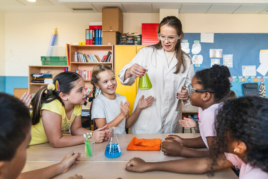 Female teacher in lab coat and safety glasses showing chemical reaction to diverse elementary students during science class
