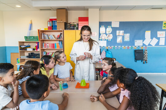 Female teacher wearing a lab coat and safety glasses engaging children with a science experiment in elementary school classroom