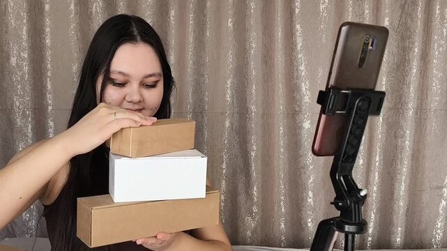 Young Asian woman holding product boxes while filming herself on camera, creating unboxing UGC content for brand promotion and social media marketing.