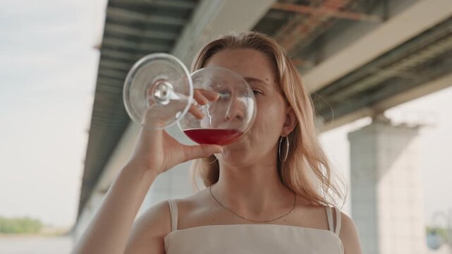 Woman holds and drinks from large wine glass under overpass with industrial structures in background. Woman wears white top and necklace interacting naturally with drink against soft