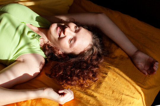 Teenage girl or young woman lying on a bed and laughing in warm sunlight. Youthful relaxation and carefree happiness. Adolescent lifestyle and personal wellbeing. Candid at-home portrait.