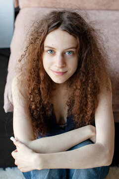 Teenage girl seated with arms around knees indoors. Close-up youth portrait. Adolescence and self expression. Individuality and contemporary youth culture. Coming of age.